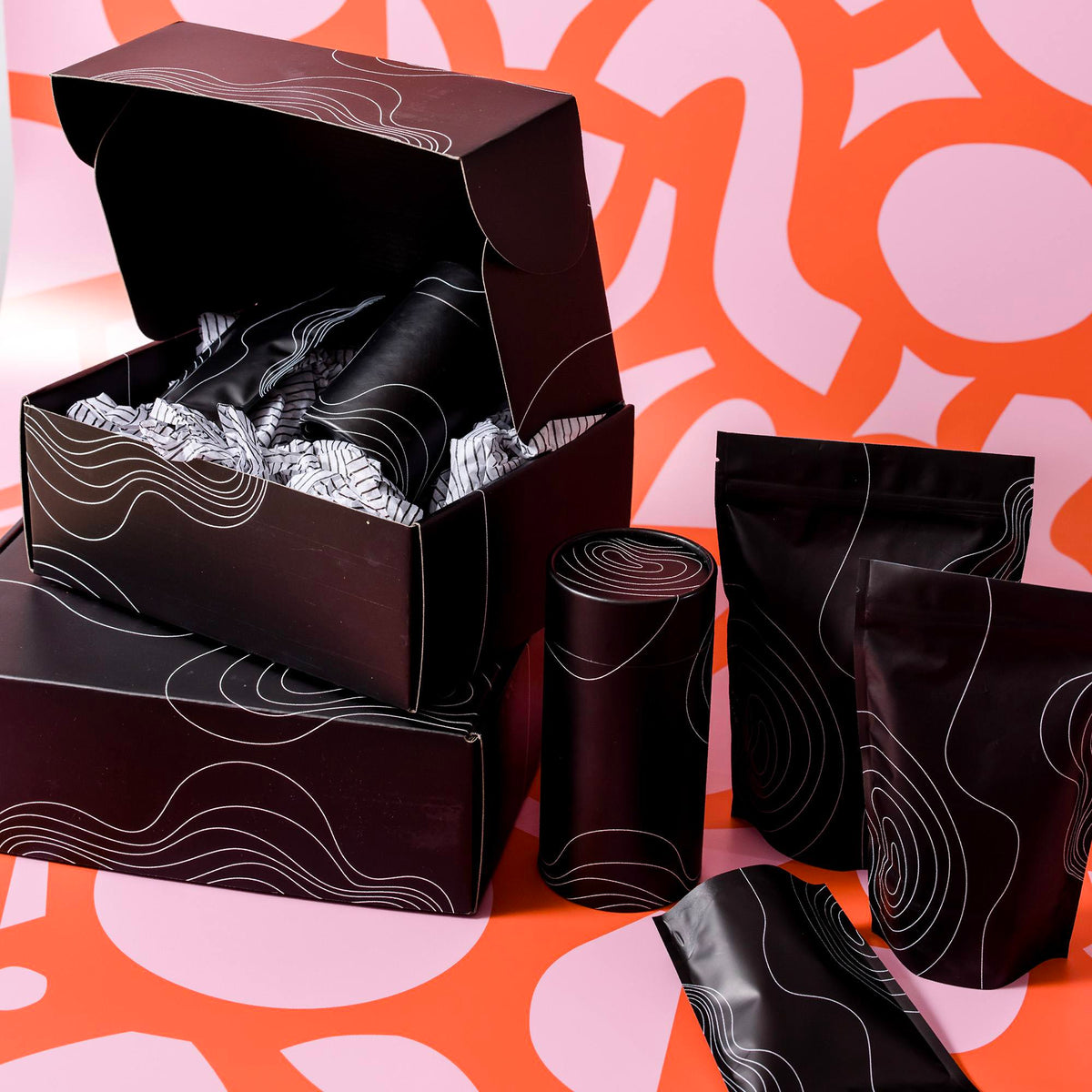 Black mailer boxes, a cylinder and stand-up pouches printed with white contour lines on a pink and orange backdrop