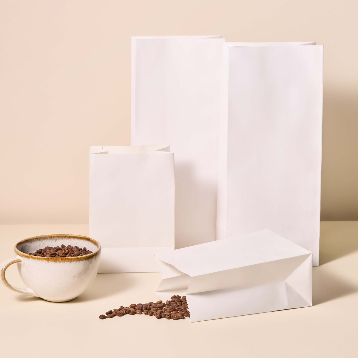 Plain white paper bags in various sizes with coffee beans spilling out beside a ceramic cup on a beige background