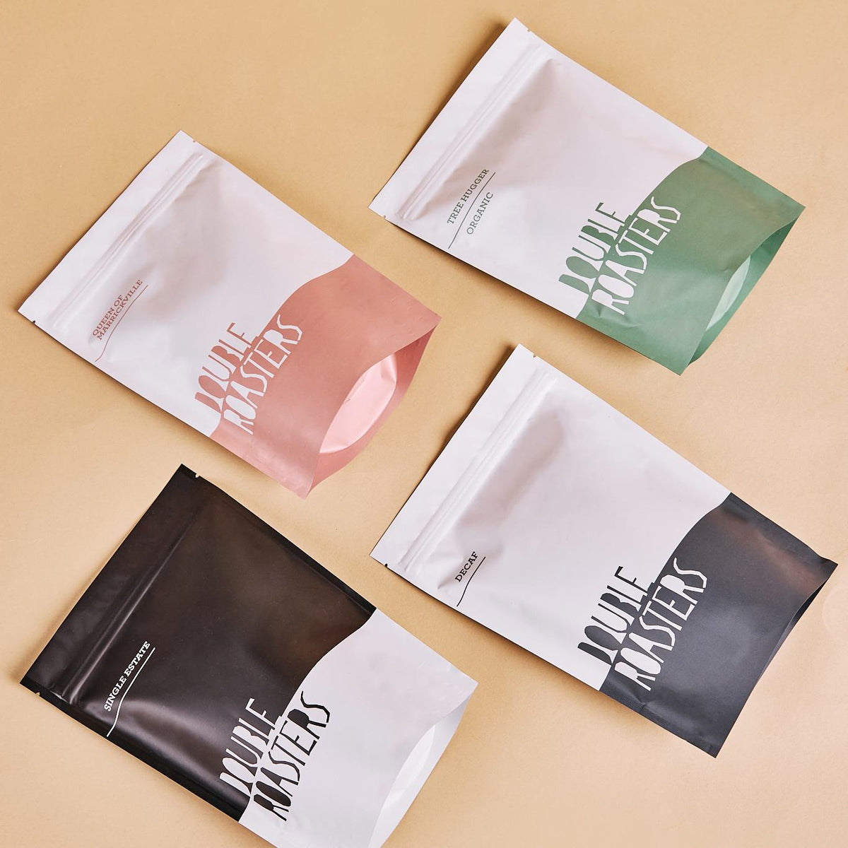 Four two-tone matte stand-up pouches with printed text, shown in pink, green, and black on a beige surface