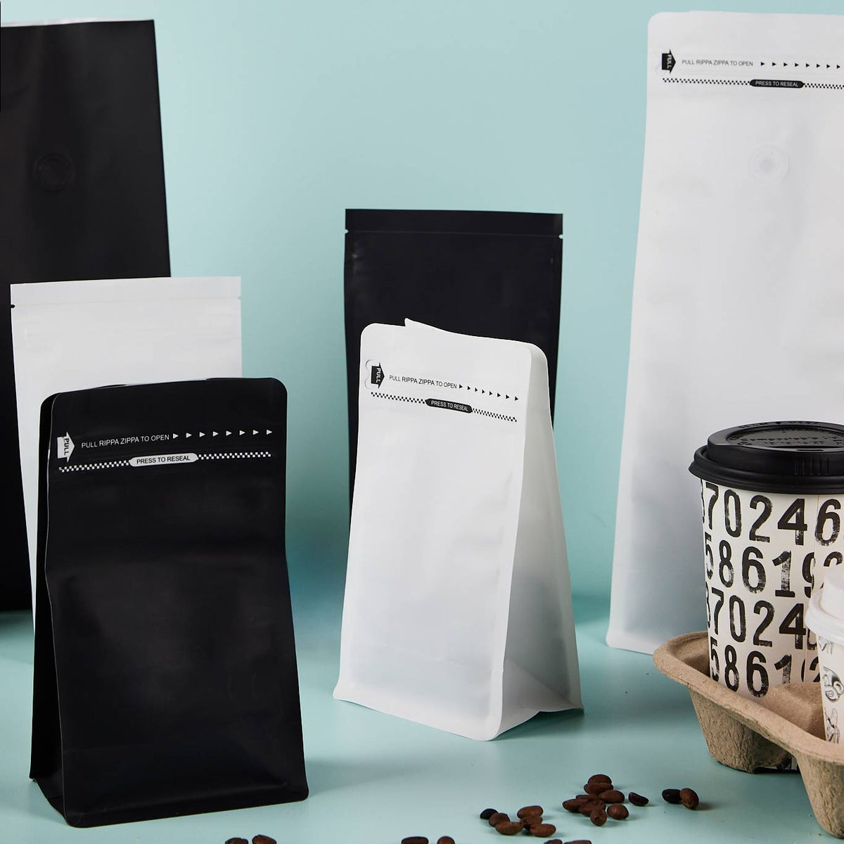 Matte black and white resealable coffee pouches on a light blue surface with scattered coffee beans and takeaway cups