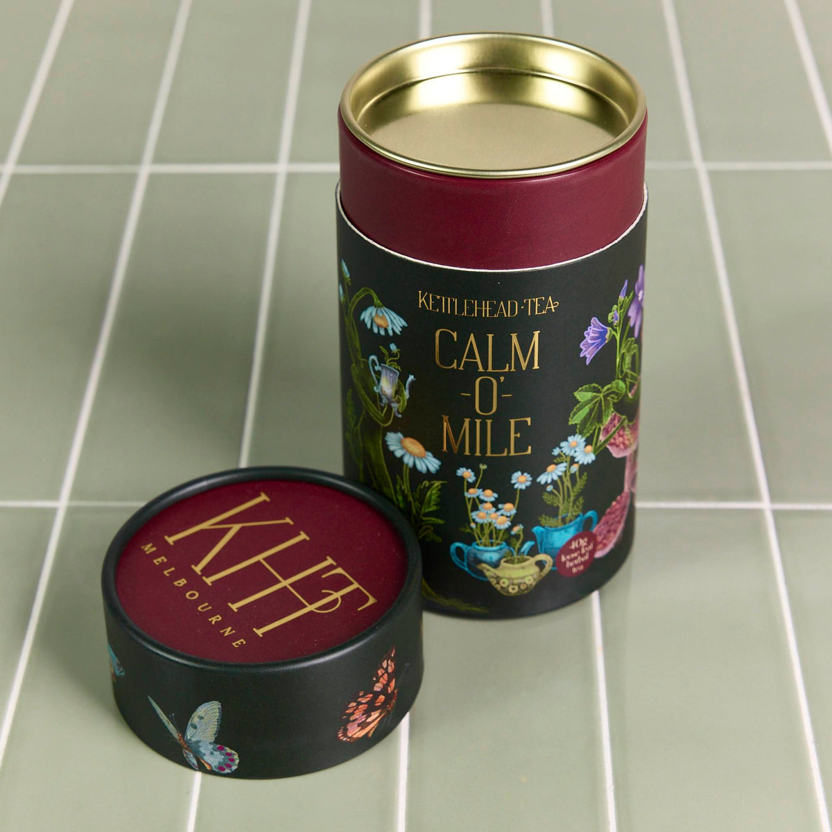Black and maroon printed tea cylinder with botanical illustrations and gold lid on a green tiled surface