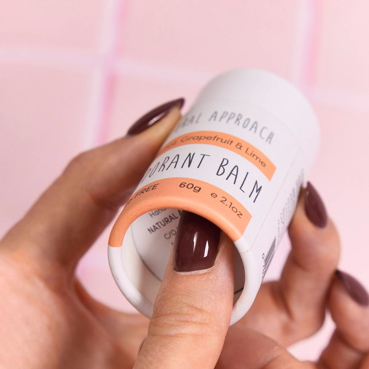 Hand pushing up the base of a white cardboard balm tube printed in black and peach, set against pink tiles