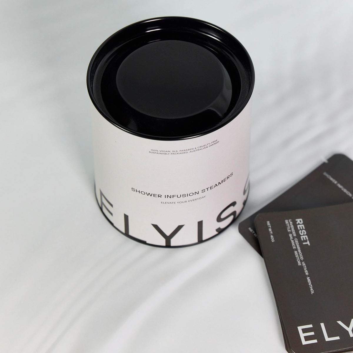 White packaging cylinder with a black lid and printed text, sitting beside black sachets on draped white fabric