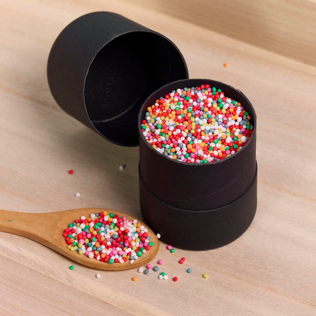 Matte black packaging cylinder filled with colourful sprinkles, beside its lid and a wooden spoon on timber