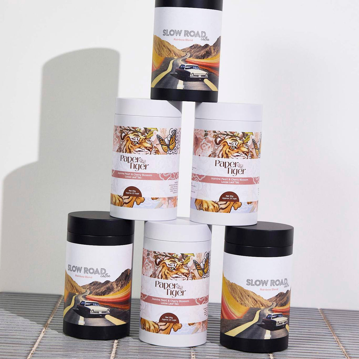 Stacked black and white packaging cylinders with custom printed tea and coffee labels on a slatted mat