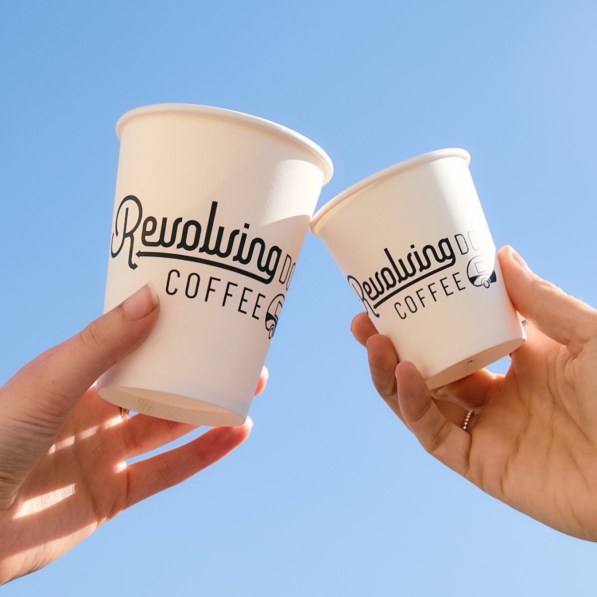 Two white takeaway coffee cups with black branding held up against a clear blue sky