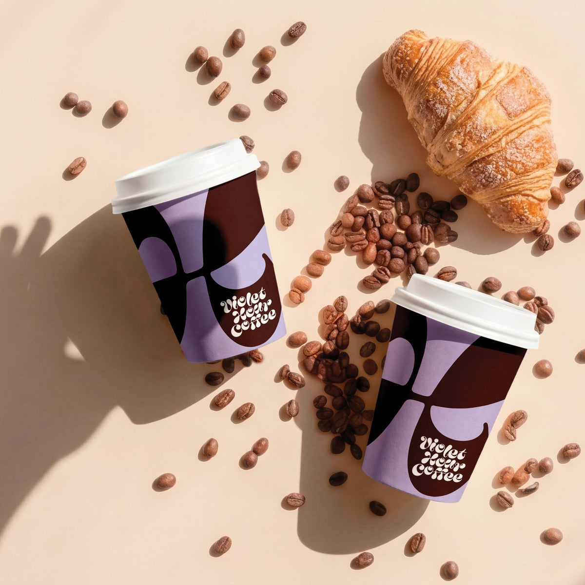 Two purple and brown printed coffee cups with white lids, surrounded by scattered coffee beans and a croissant