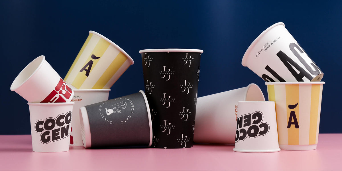 Custom-printed paper coffee cups in assorted sizes and designs arranged on a pink surface against a blue backdrop