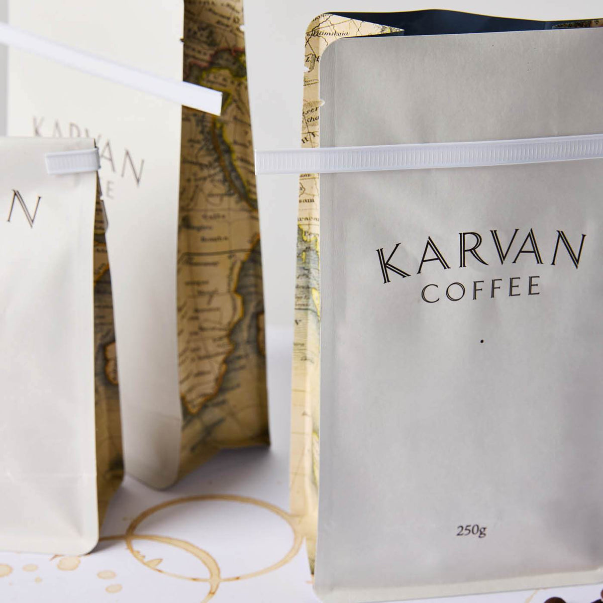 Matte white Karvan Coffee bags with map-printed gussets and white tin ties on a surface with coffee rings