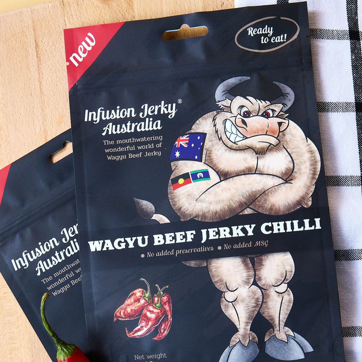 Matte black stand-up pouch with a hang hole and printed jerky design, on a timber board beside a fresh red chilli