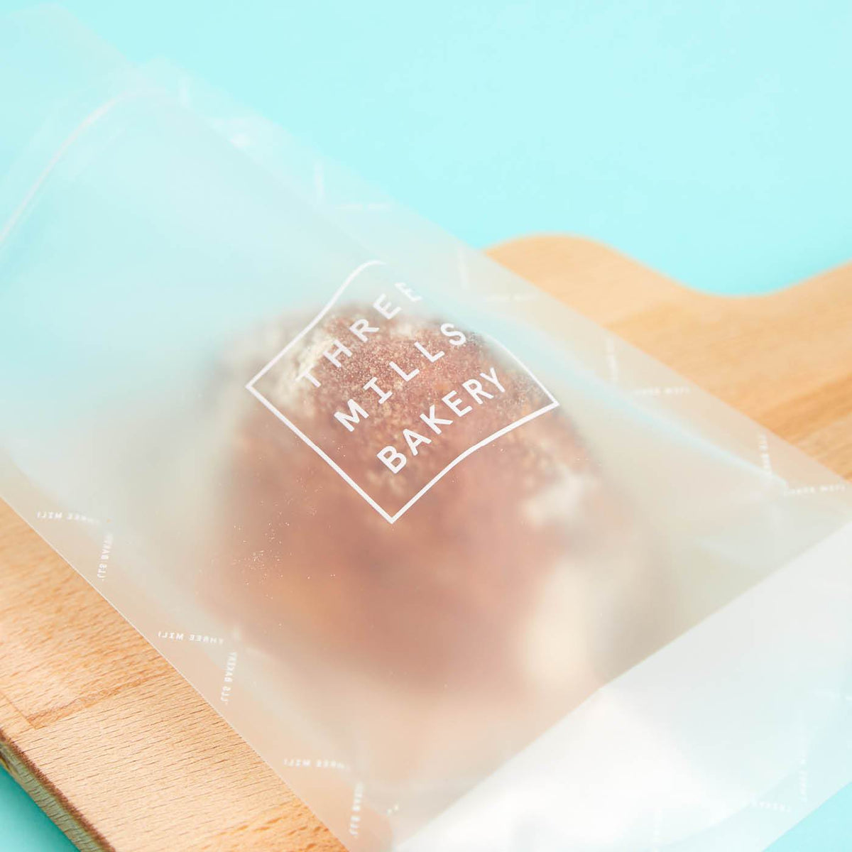 Frosted stand-up pouch printed with a white Three Mills Bakery logo, holding a pastry on a wooden board