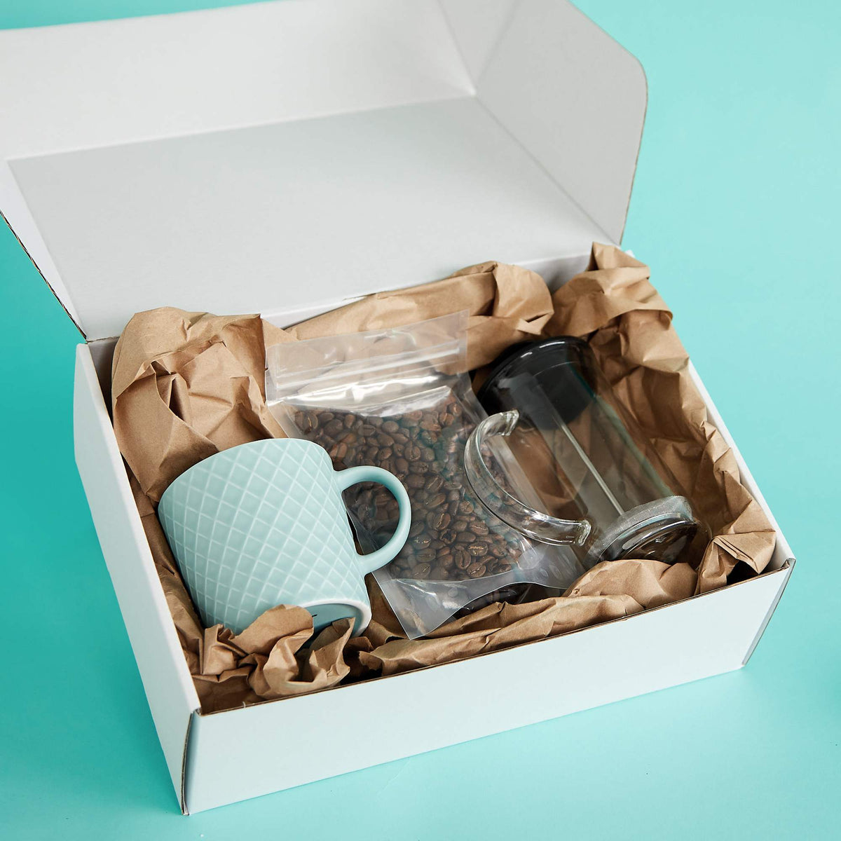Open white mailer box packed with kraft paper, a blue mug, coffee beans, and a glass plunger on a turquoise background