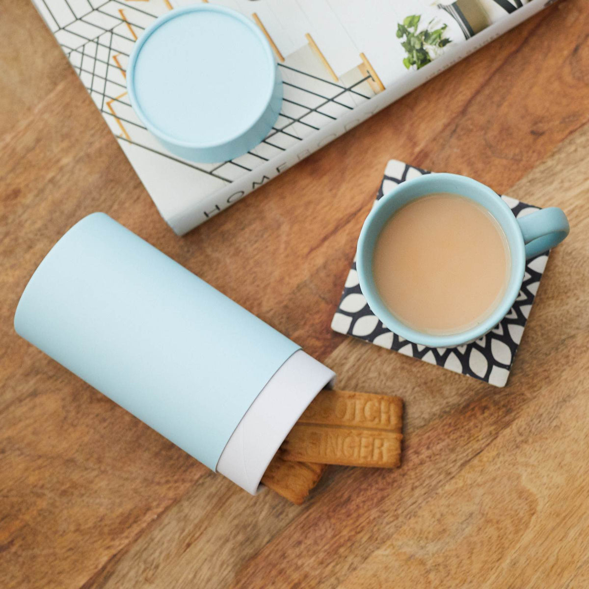Open light blue cardboard packaging cylinder with biscuits inside, next to a matching cup of tea on a wooden table