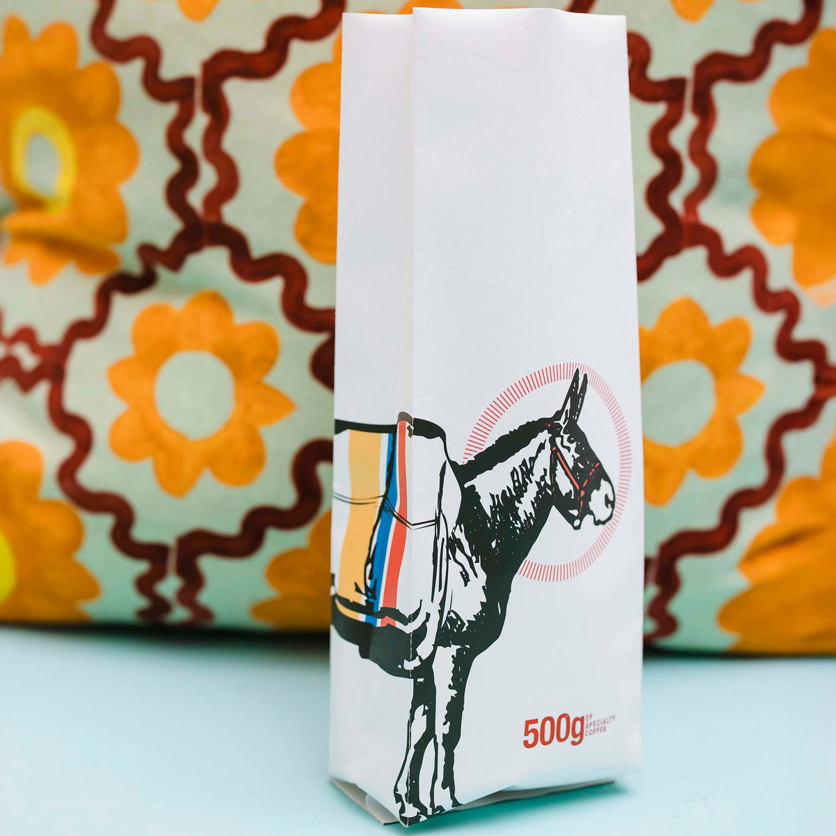 White side-gusset coffee bag printed with a donkey illustration and 500g text, against a patterned cushion