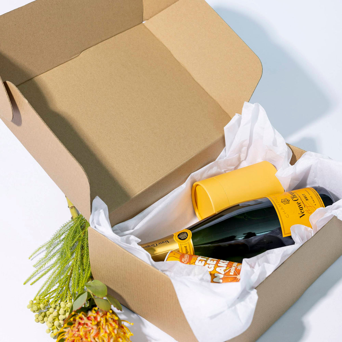 Open brown kraft mailer box lined with white tissue paper, holding a champagne bottle and a yellow cardboard cylinder