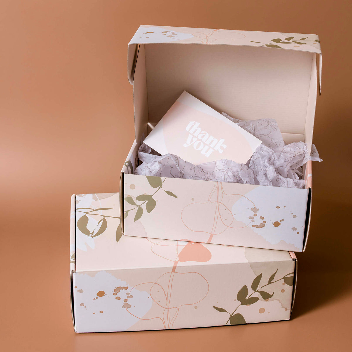 Two cream mailer boxes with a botanical print on a tan background, one open with tissue paper and a card