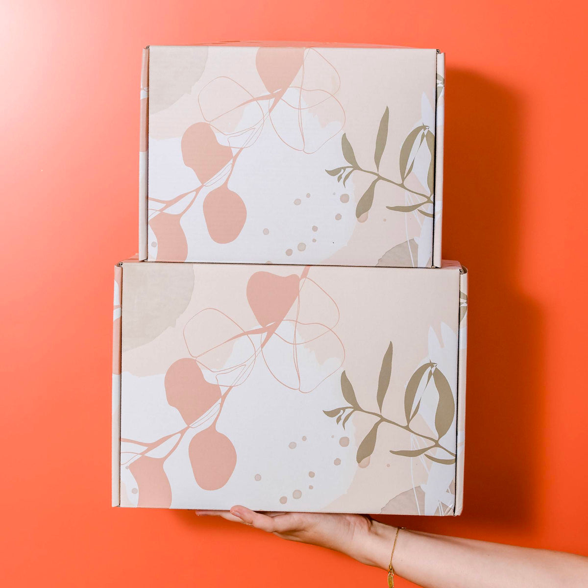 Two stacked mailer boxes printed with beige botanical artwork, held against a terracotta-coloured background