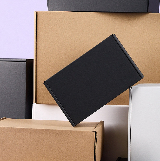 Matte black mailer box balanced diagonally against stacked plain kraft and white mailer boxes