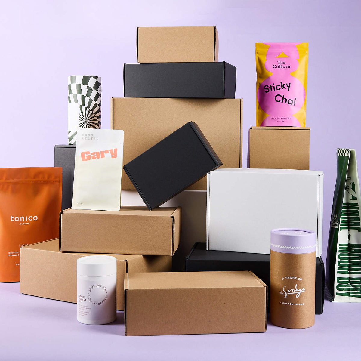 Stacked kraft, black and white mailer boxes with colourful printed pouches, cylinders and a jar on a lilac background