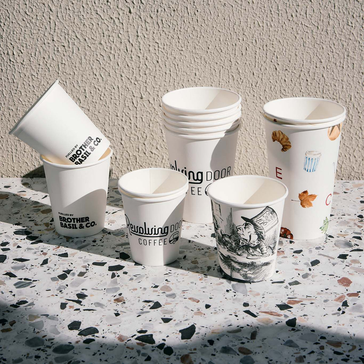 Custom-printed white paper coffee cups in various sizes arranged on a terrazzo surface against a textured wall