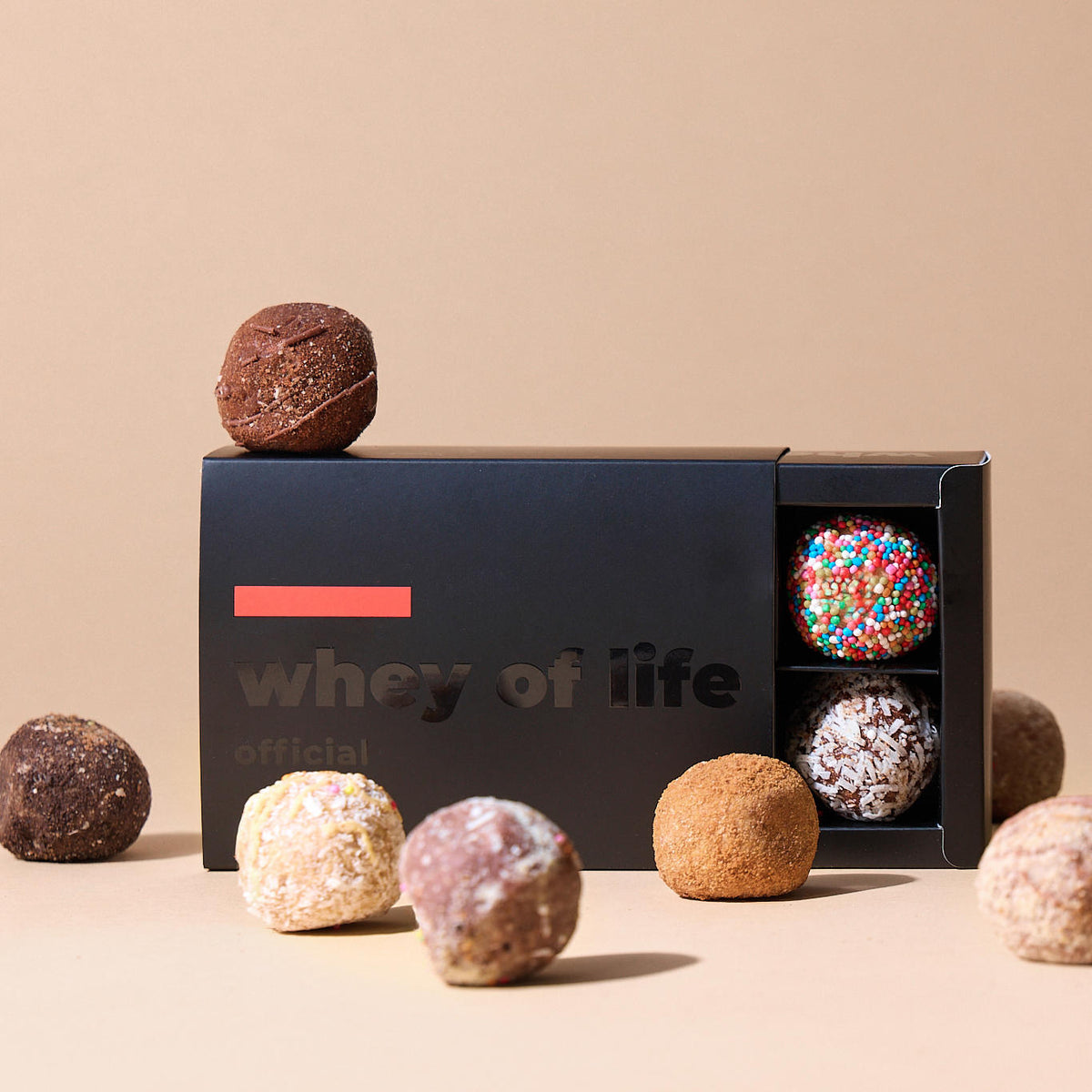 Matte black sliding box printed with whey of life, holding assorted snack balls on a beige surface