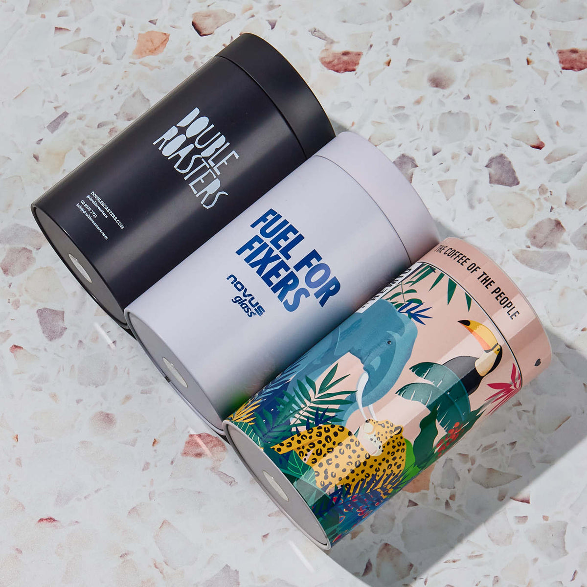 Three printed packaging cylinders in matte black, light grey, and colourful jungle artwork on a terrazzo surface