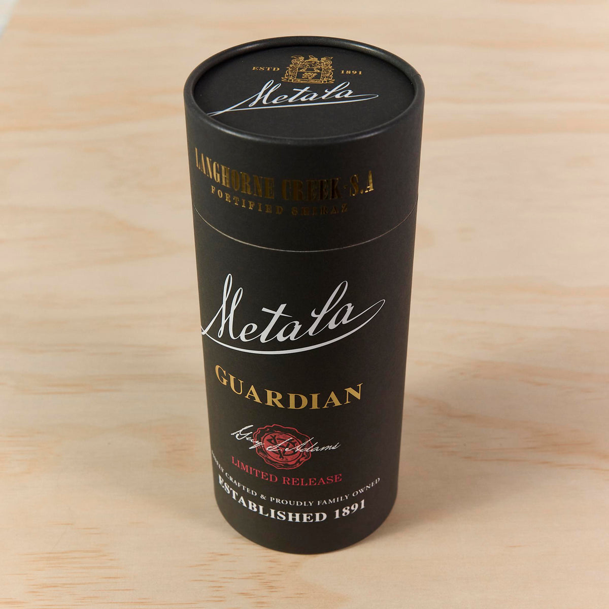 Matte black packaging cylinder with custom gold and white print, standing on a light timber surface