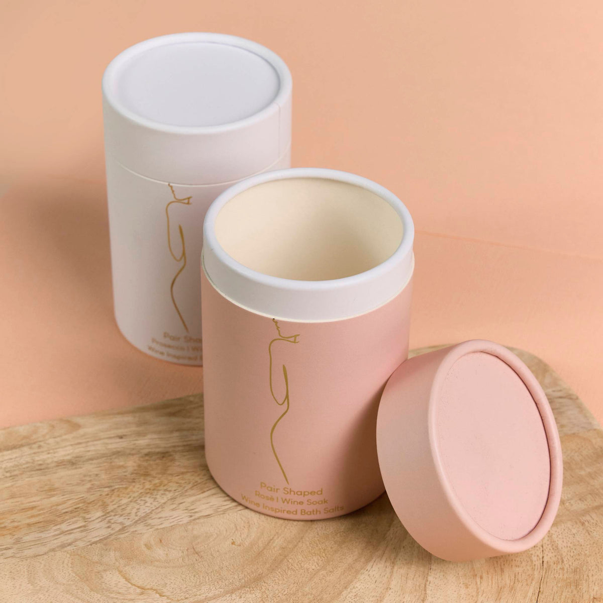 Matte pink and white packaging cylinders with gold line art, displayed open and closed on a wooden board