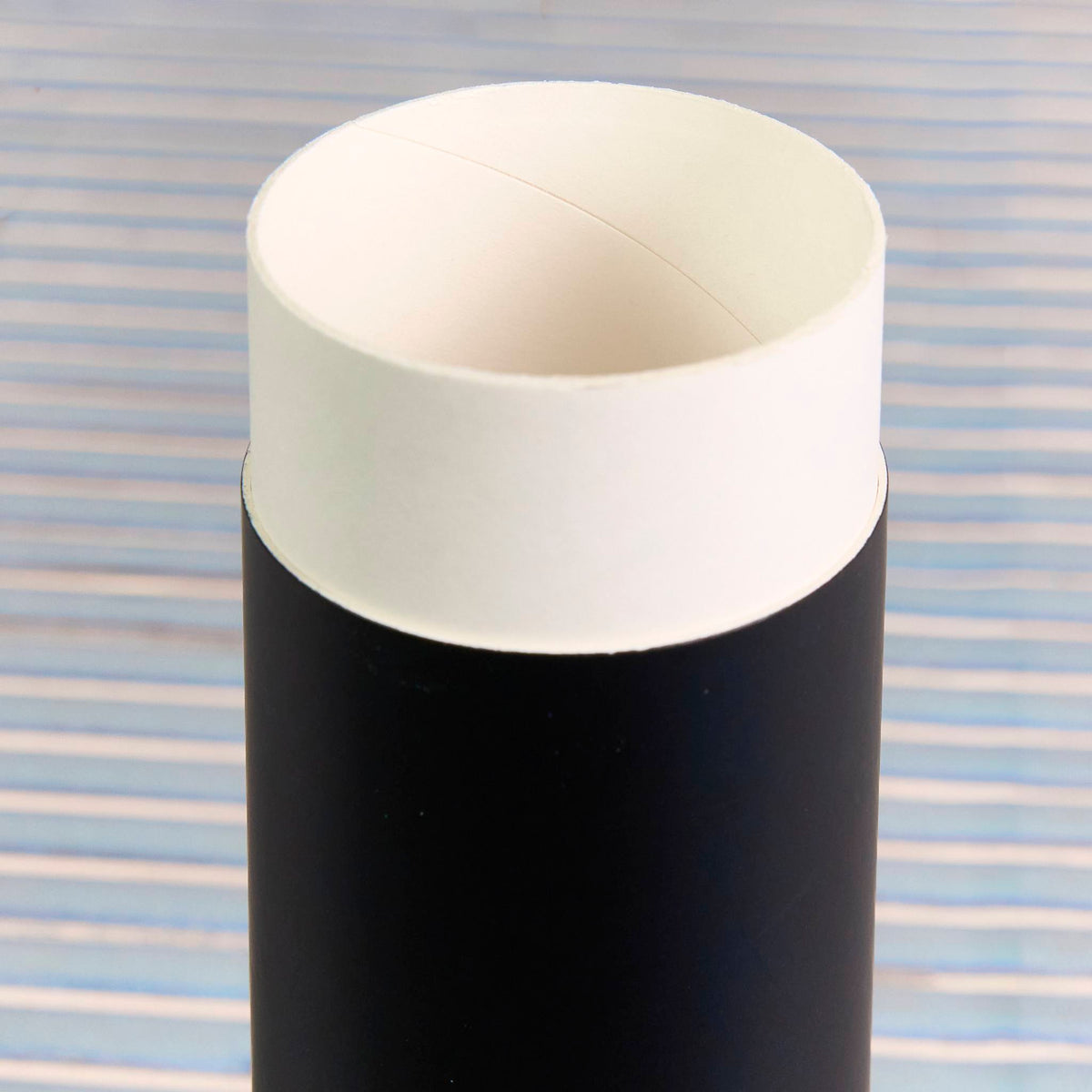 Open matte black cardboard cylinder with a white inner lip, shown against a light blue striped background