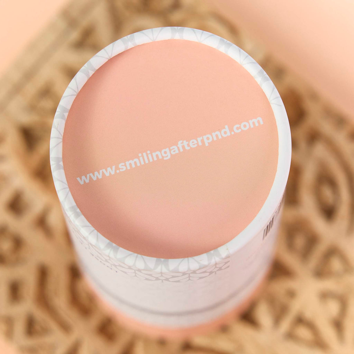 Top view of a cardboard packaging cylinder with a peach lid and patterned rim, resting on a carved wooden surface
