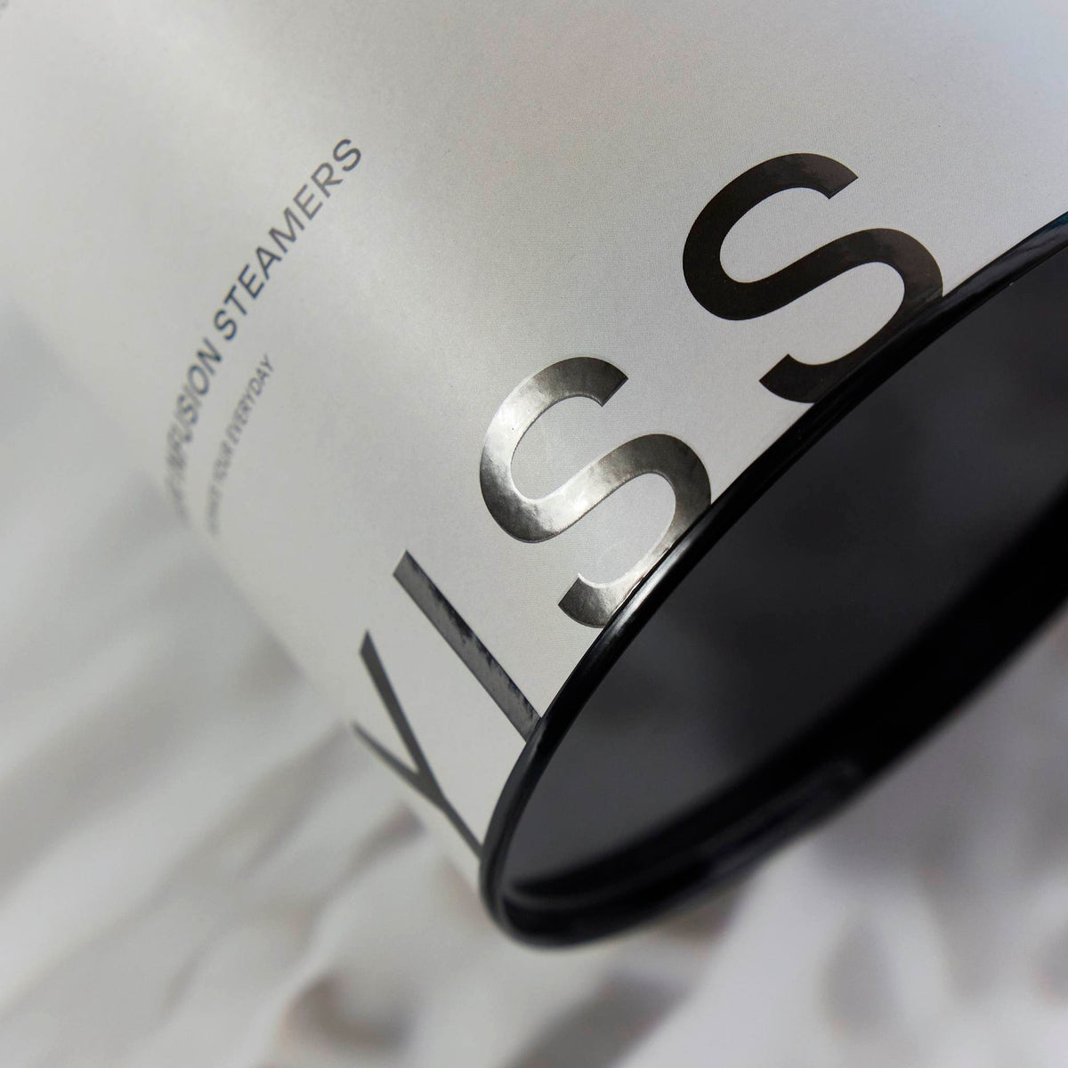 Close-up of a white packaging cylinder with metallic foil lettering and a black base, resting on white fabric