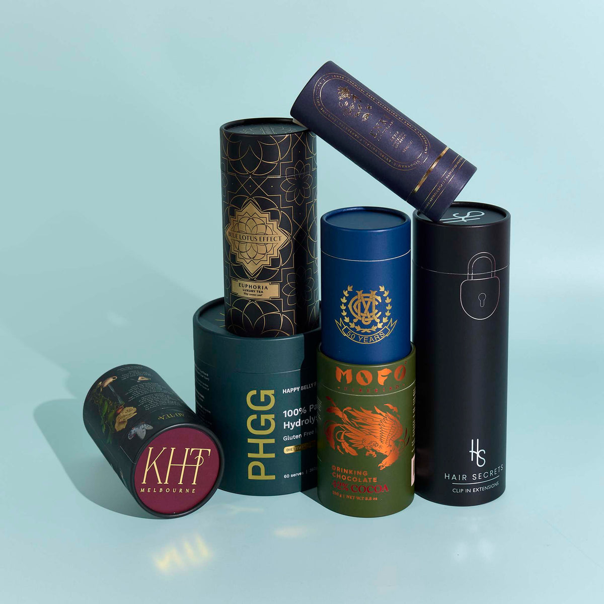 Assorted matte packaging cylinders with custom metallic foil print in various colours on a light blue background