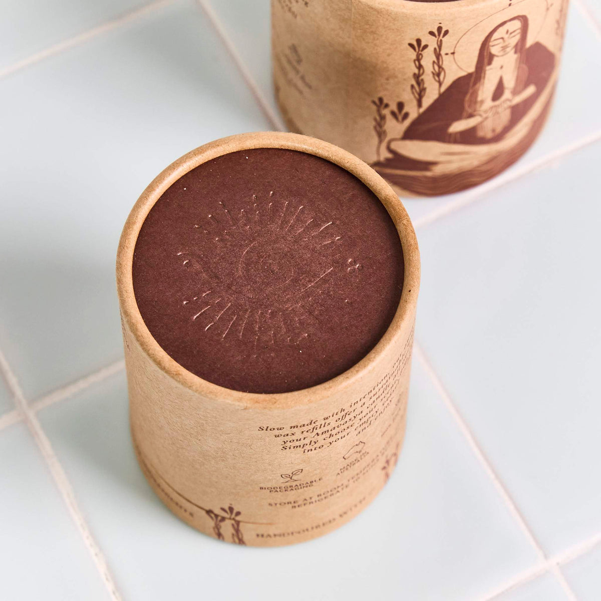 Kraft packaging cylinder with printed text and an embossed dark brown lid on light blue tiles