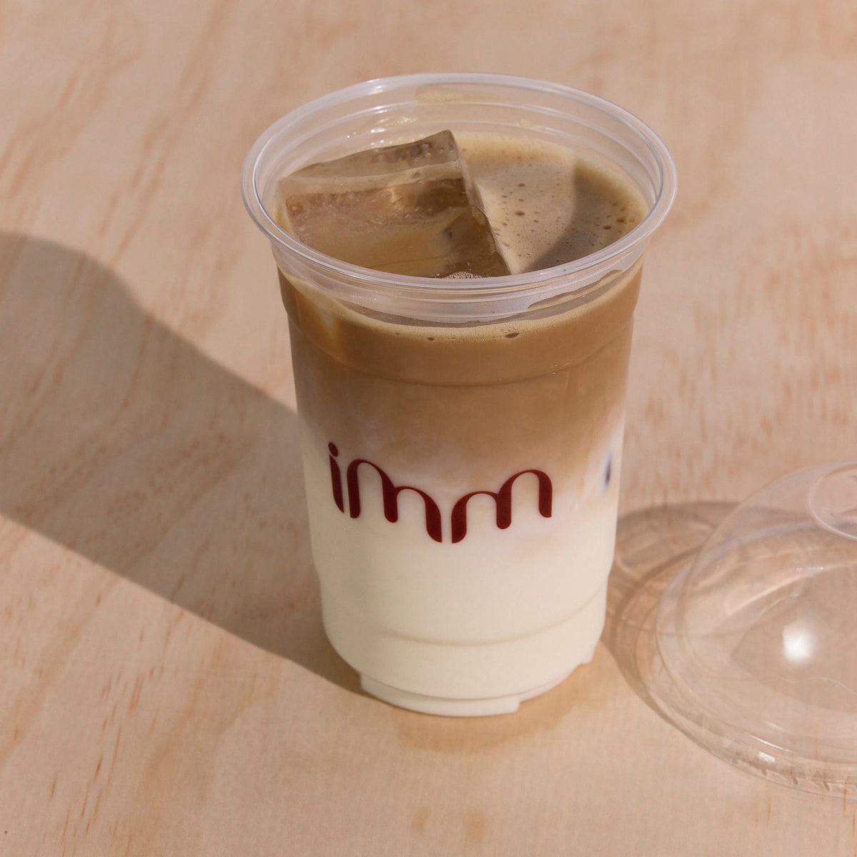Clear plastic cold cup printed with maroon "imm" lettering, holding iced coffee on a timber surface beside a dome lid