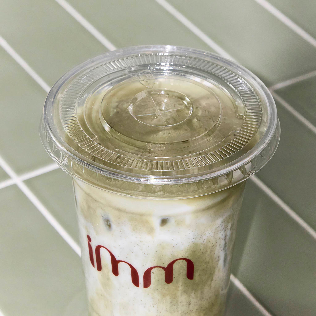 Clear plastic cold cup with a flat lid and red printed logo, filled with an iced drink on a green tiled surface