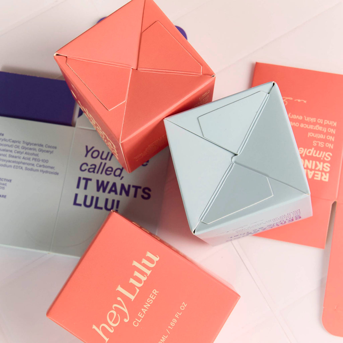 Custom printed folding cartons in coral pink and pale blue, showing interlocking base folds on a flat-lay surface