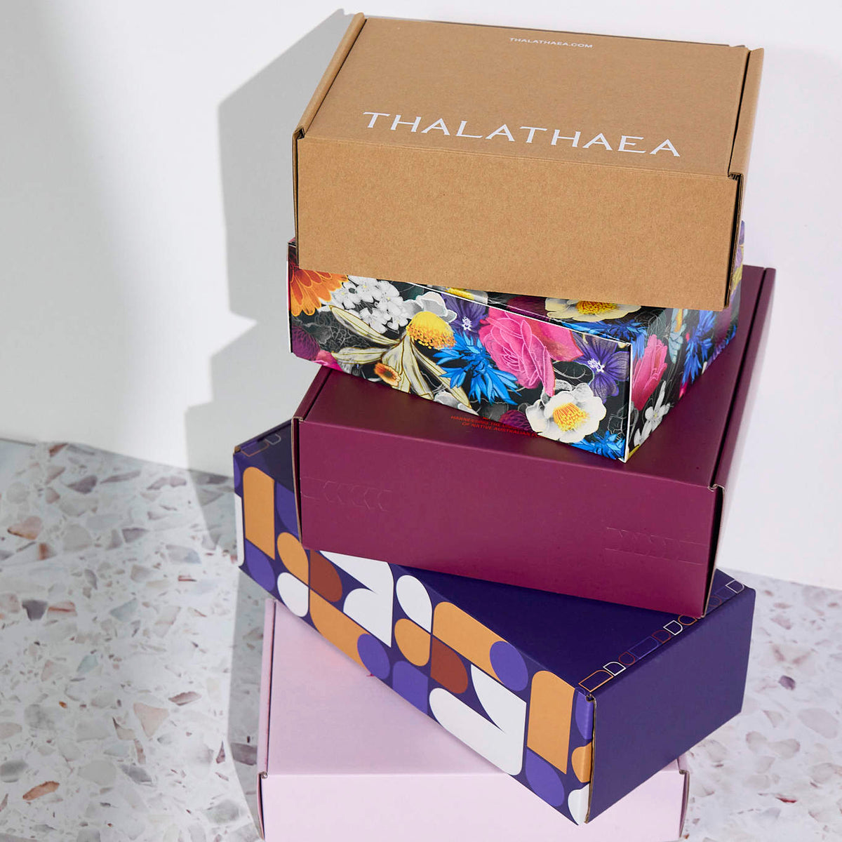 Stacked custom mailer boxes in kraft, floral, geometric, and solid colours on a terrazzo surface