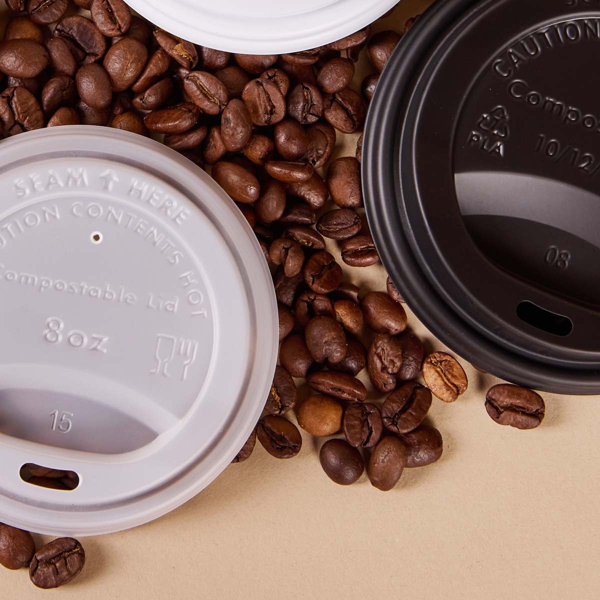 White and black compostable coffee cup lids displayed over roasted coffee beans on a beige background