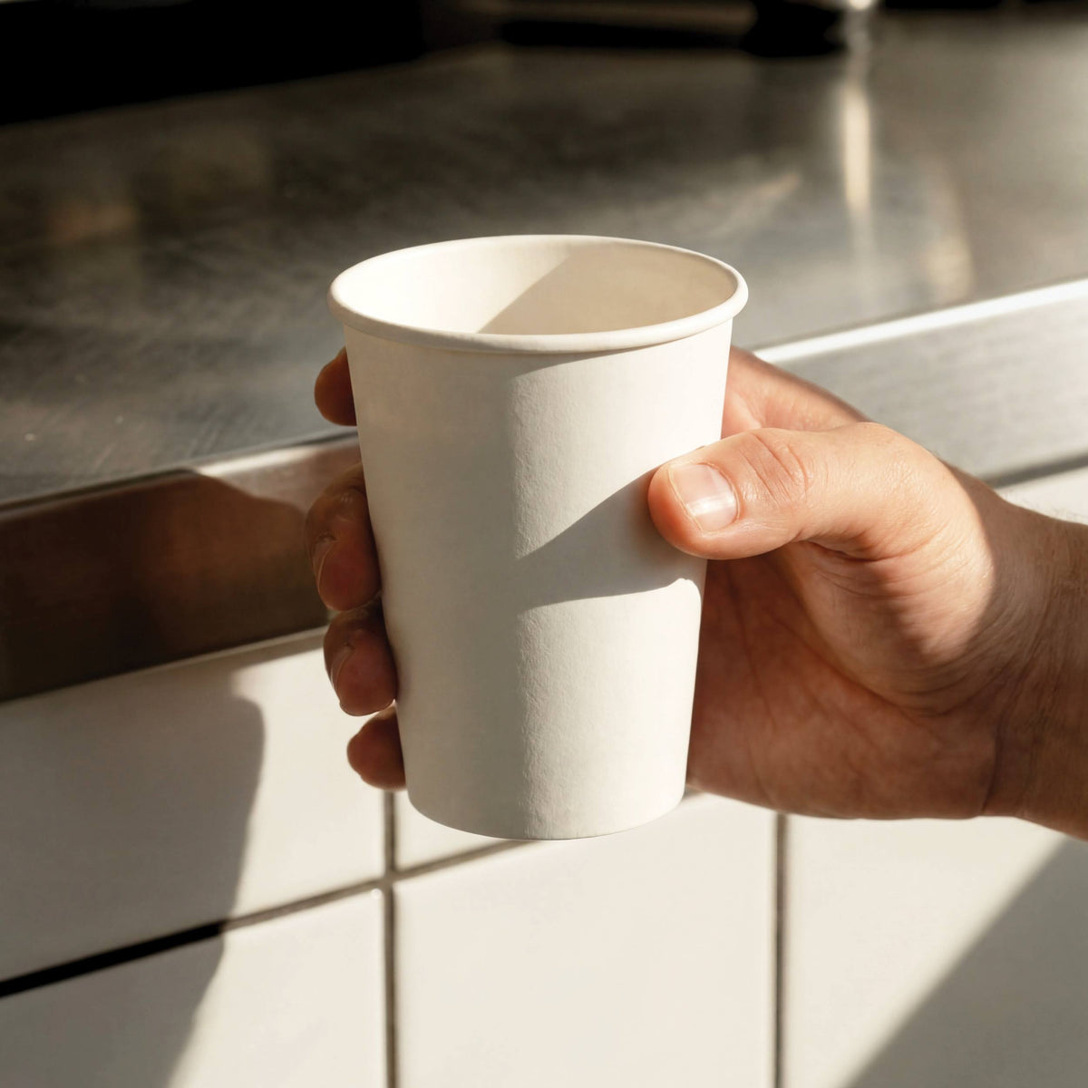 A hand holding a plain white paper coffee cup by a stainless steel counter