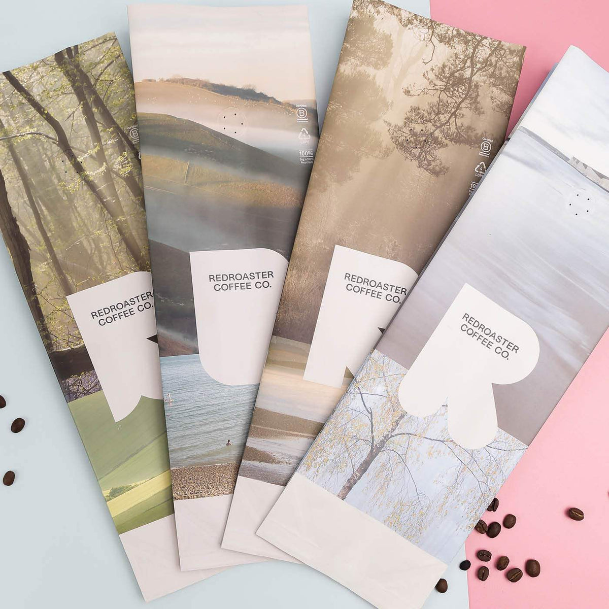 Four matte coffee bags printed with landscape photos, displayed on a pink and blue surface with scattered coffee beans