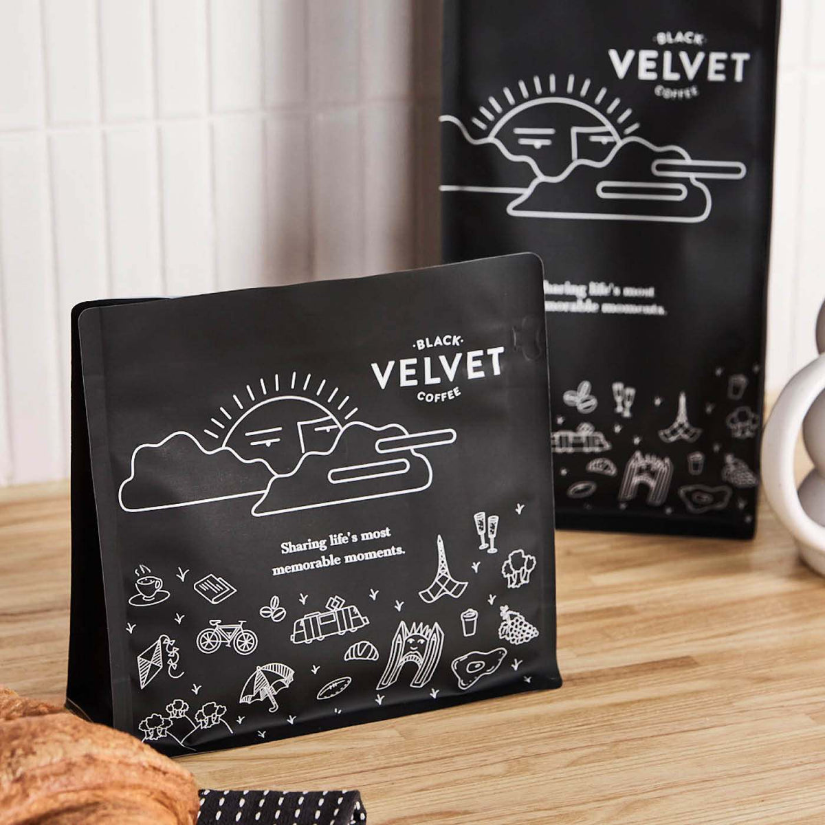 Matte black stand-up pouch printed with white line illustrations, on a timber benchtop against white tiles