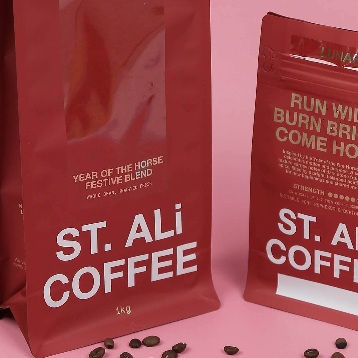 Matte red stand-up coffee pouches printed with white text, on a pink surface with scattered coffee beans