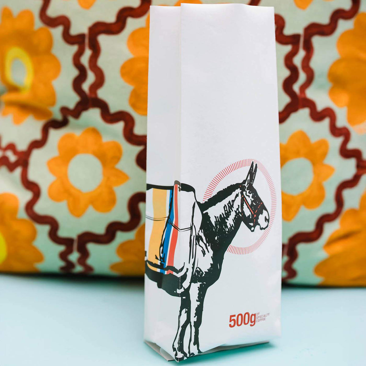 Matte white side-gusset coffee bag printed with a donkey illustration, set against a patterned cushion