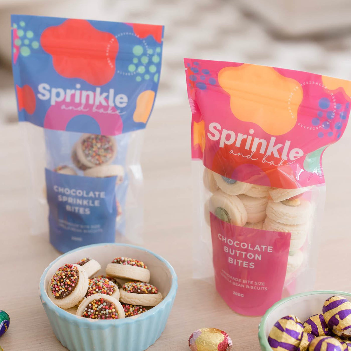 Two colourful, glossy stand-up pouches with clear windows holding biscuits, displayed on a table beside small bowls