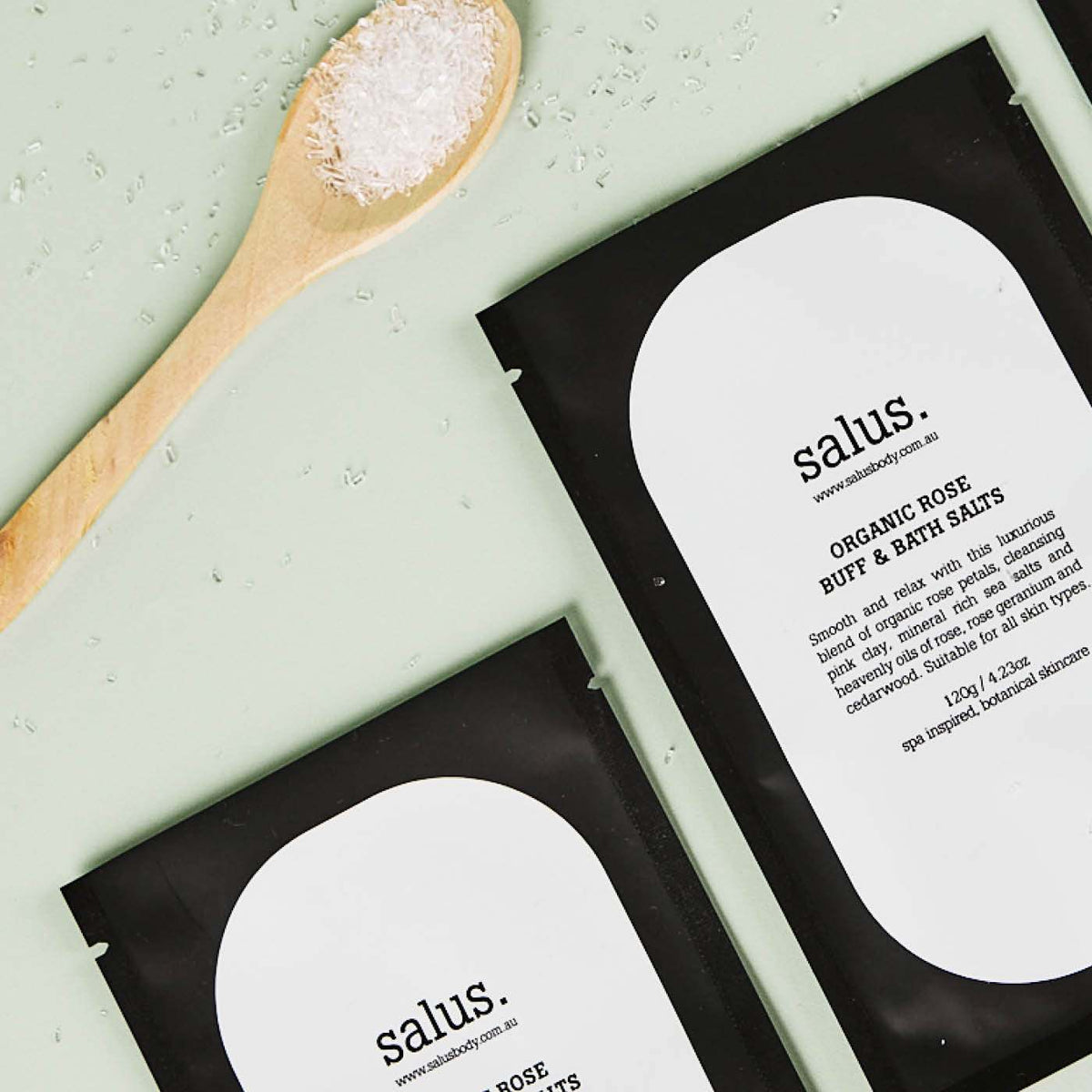 Black stand-up pouches with a white arch design, beside a wooden spoon filled with bath salts on a pale green surface