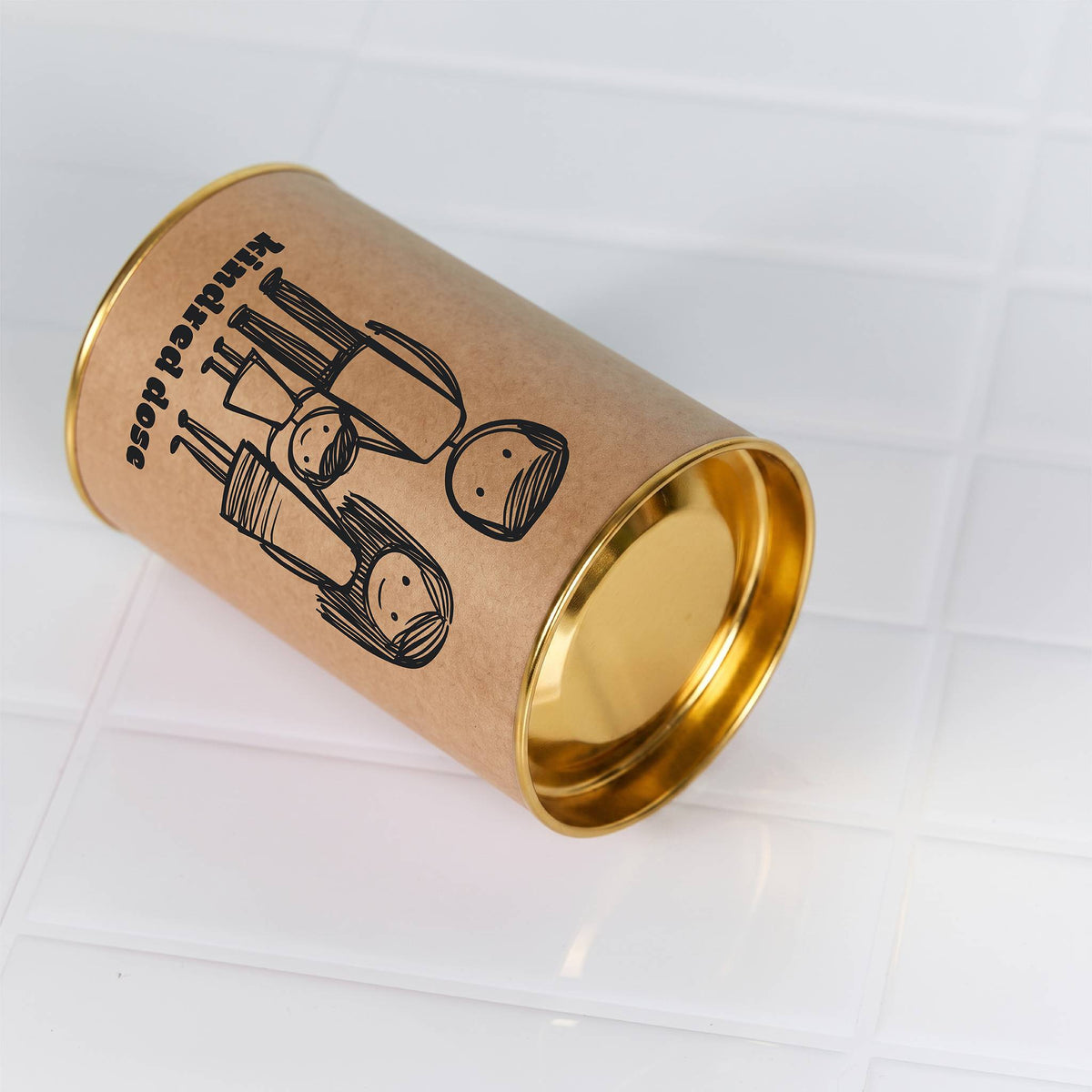 Kraft paper cylinder with gold metal ends and a black printed illustration, resting on a white tiled surface
