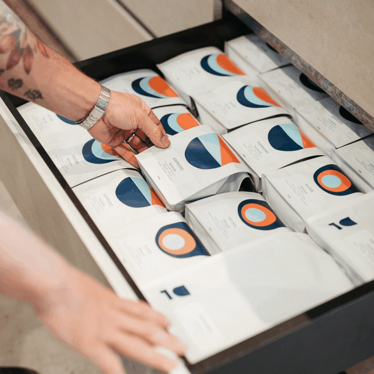 A hand reaches into an open drawer filled with white coffee pouches featuring blue and orange geometric designs