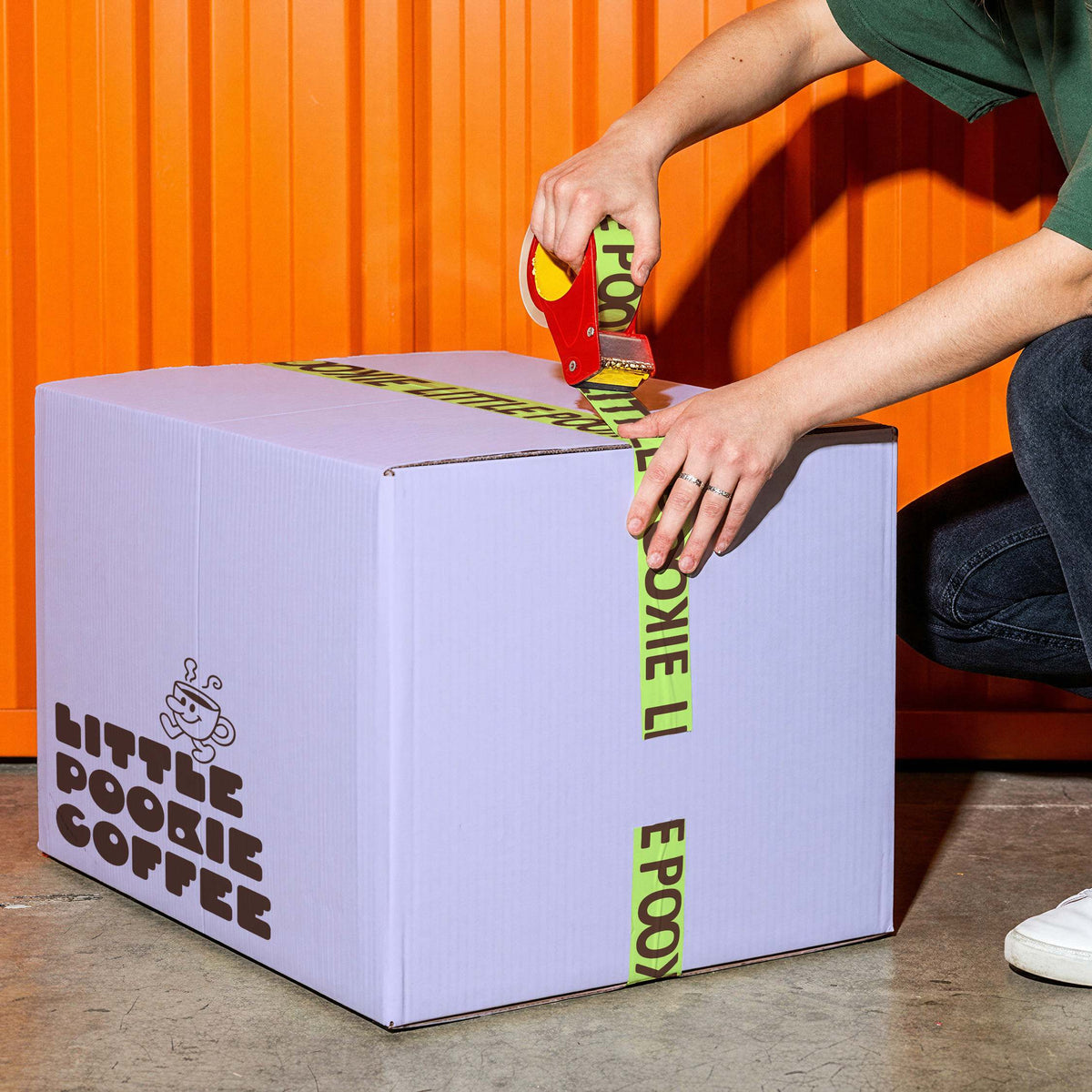Lilac shipping carton printed with a black coffee logo, being sealed with green packing tape against an orange wall