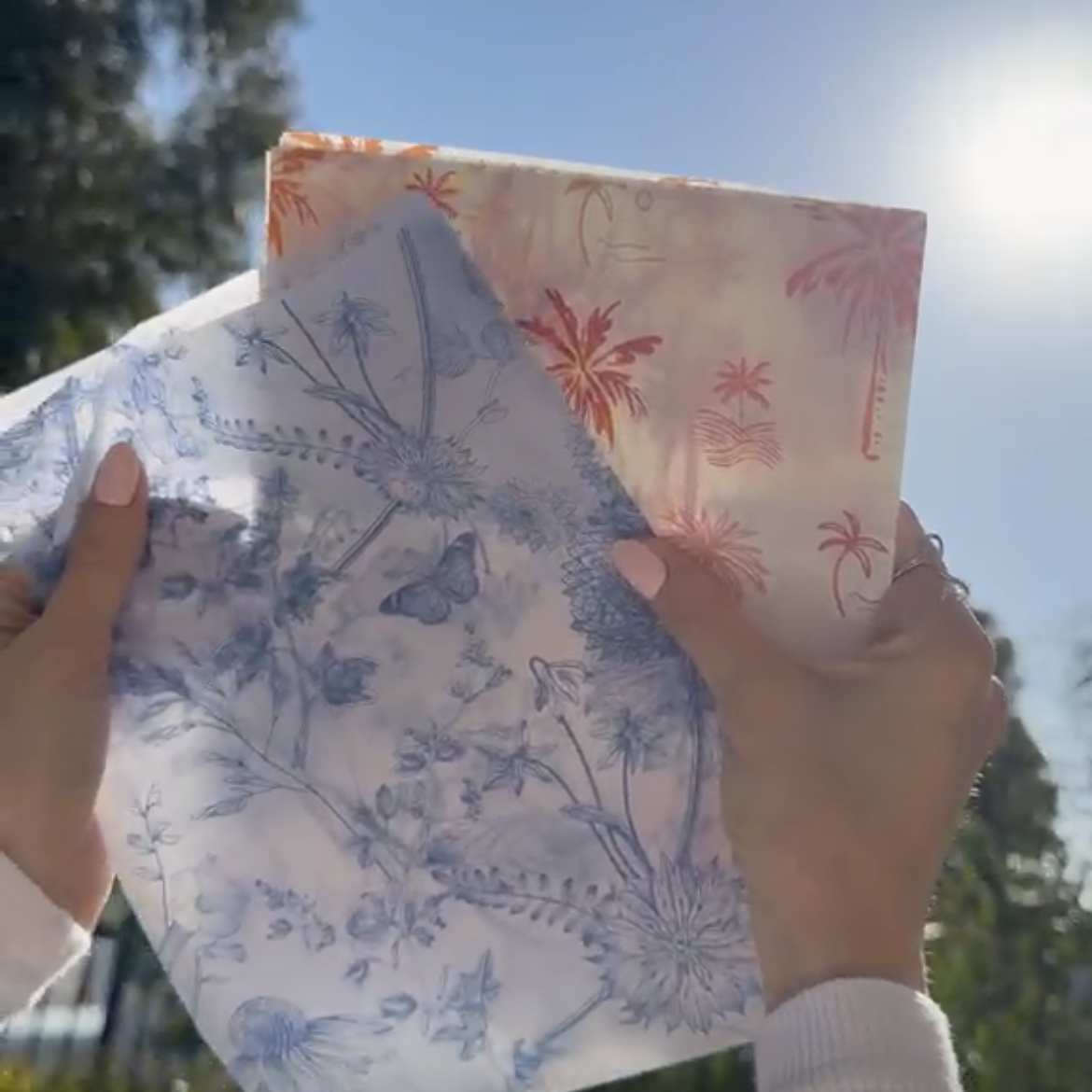 Hands holding custom tissue paper sheets printed with blue floral and pink palm tree designs against a sunny sky